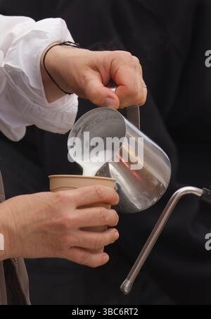 Primo piano di un barista che versa latte appena montato in una tazza, preparando una bevanda calda con cura e precisione. Immagine ideale per la preparazione del caffè Foto Stock