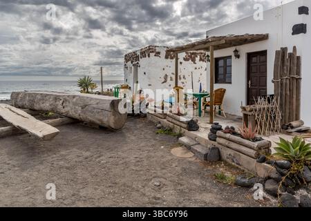 Un tavolo e sedie di legno si trovano in un giardino rustico davanti a una piccola casa bianca a El Golfo sull'Avenida Marítima, El Golfo, Lanzarote, Canarie, Spagna. Foto Stock