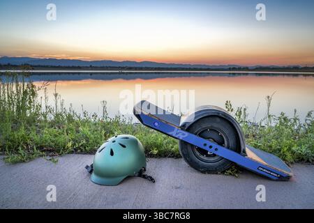 Fort Collins, CO, USA - 11 agosto 2018: Skateboard elettrico OneWheel con casco su un lago addolorato contro il cielo del tramonto Foto Stock