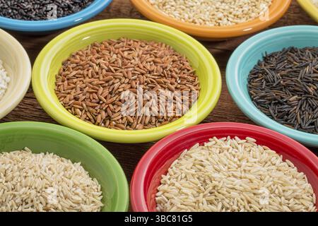 Una varietà di grani di riso su ciotole di ceramica colorata contro un legno a grana Foto Stock