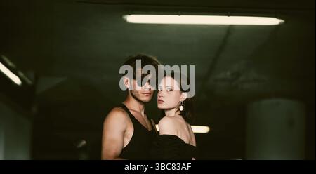 Una coppia elegante con un abbigliamento scuro si posa sotto una plafoniera in un ambiente poco luminoso Foto Stock