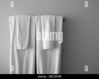 Coppie di asciugamani bianchi puliti, grandi e piccoli, piegati appesi a una barra di appendiabiti in metallo con piastrelle grigie all'angolo del bagno, dietro le quinte orizzontale Foto Stock