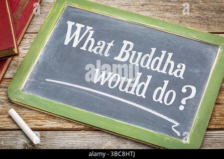 Cosa farebbe Buddha? Una domanda ispiratrice su una lavagna in ardesia con gesso bianco e una pila di libri su un tavolo in legno rustico Foto Stock