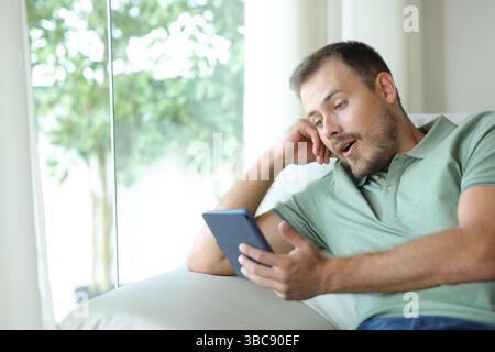 Uomo sorpreso che leggesse un eBook seduto su un divano in una casa al coperto Foto Stock