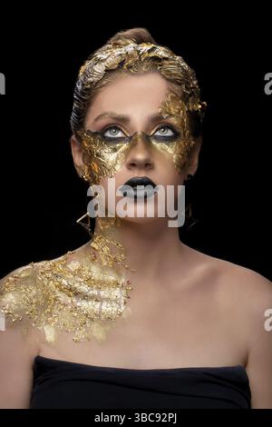 Primo piano di una bella giovane donna con un foglio dorato sul viso. Trucco creativo dorato su sfondo nero Foto Stock