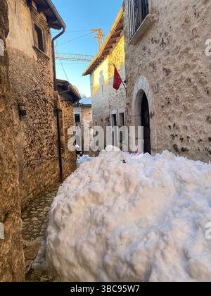 Stretta strada innevata che si snoda tra storiche abitazioni in pietra a Santo Stefano di Sessanio, in Abruzzo, con gru da costruzione che si innalza dietro la media Foto Stock