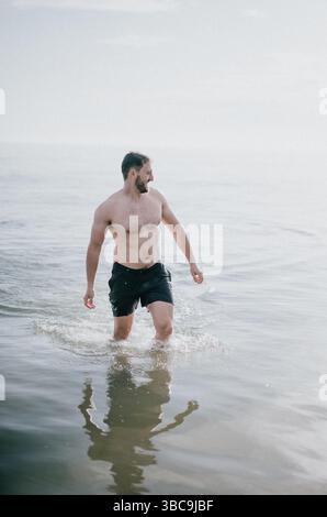 Un uomo sta camminando nell'oceano indossando solo un costume da bagno nero. Sta sorridendo e si sta divertendo Foto Stock