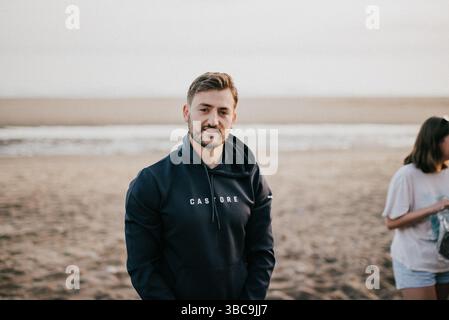 Un uomo è in piedi su una spiaggia con indosso una felpa con cappuccio con le lettere "auto". Sta sorridendo e guardando la telecamera Foto Stock