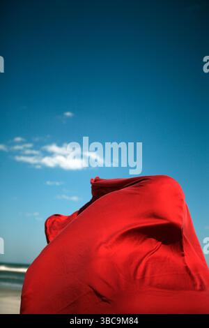 Una donna avvolta in un panno rosso si erge su una spiaggia ventosa Foto Stock