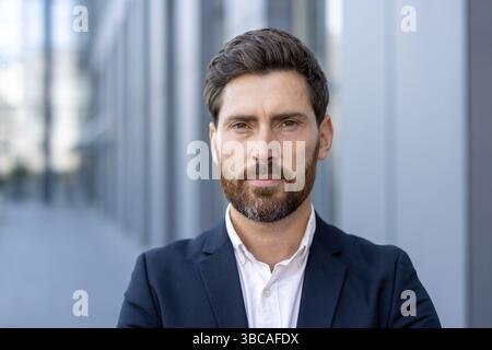 Un uomo d'affari ben curato con una barba e un'espressione seria si erge all'aperto, contro un edificio sfocato. Foto Stock