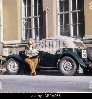 Una donna vicino a un'auto sportiva. La vettura è un modello MG TD inglese, prodotto tra il 1950 e il 1953. Questo modello era particolarmente adatto per i clienti svedesi in quanto poteva essere costruito con il volante sul lato sinistro, in linea con le convenzioni di guida svedesi dell'epoca. Sono state prodotte in totale 29.664 unità dell'MG TD. È seduta sulla macchina, a leggere la rivista femminile Femina. È vestita con un blazer e pantaloni marroni. Svezia degli anni '1960. Ulteriori informazioni sulla MG TD la MG TD, parte della serie MG T-Type, era una roadster a due posti rinomata per il suo classico design britannico, Feat Foto Stock