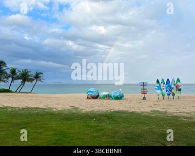 Una tranquilla spiaggia tropicale in Vietnam e' incorniciata da palme e tavole da surf colorate, mentre un debole arcobaleno si arrocca sul mare calmo sotto un cielo morbido e nuvoloso Foto Stock