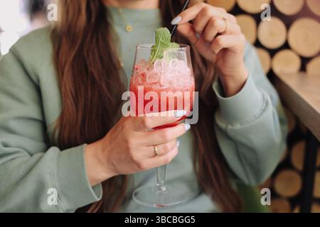 Cocktail rosso rinfrescante con ghiaccio. Donna che tiene in mano un drink. Bevanda estiva con foglie di menta. Elegante vetro con paglia. Rilassatevi nel bar. Foto Stock