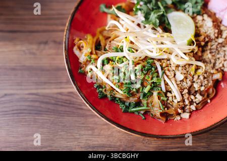 Tradizionale piatto di noodle asiatico. Colorato pad thai su piatto rosso. Erbe e verdure fresche. Arachidi croccanti e lime. Autentica cucina orientale. Foto Stock