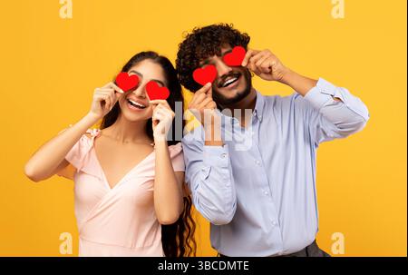 Amanti accecati dal loro grande amore. Giovane coppia indiana innamorata che tiene carte rosse a forma di cuore sugli occhi Foto Stock