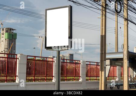 Cartellone verticale vuoto su un marciapiede vicino ai pali elettrici e alla recinzione rossa con moderni edifici cittadini sullo sfondo. Perfetto per pubblicità all'aperto Foto Stock
