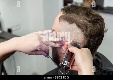 uomo con barba dai capelli scuri adulto nel moderno barbiere durante il processo di taglio dei capelli con rasoio elettrico. Utensili e accessori professionali in mano al barbiere Foto Stock