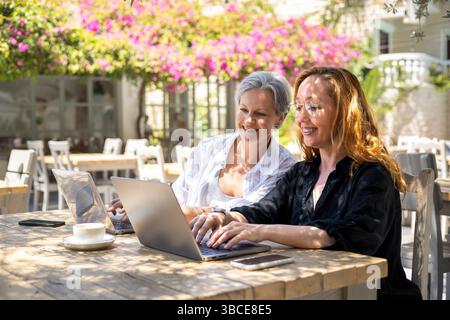 Donne mature che lavorano in remoto su notebook all'Outdoor Cafe, sorridenti e collaborano a Sunny Weather Foto Stock