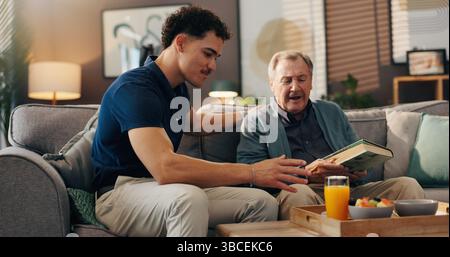 Accompagnatore, anziano e colazione sul divano, lettura o conversazione con libro, cibo o nutrizione in lounge. Infermiera maschile, paziente anziano e sorriso per il pasto Foto Stock