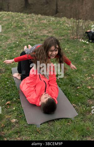 I momenti di gioia sono condivisi mentre due bambini si impegnano in pose giocose su un tappetino yoga, circondati da una vegetazione vibrante e da un senso di spensieratezza divertimento e connessione Foto Stock