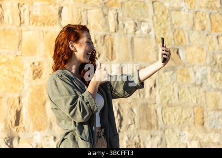 Giovane donna allegra con i capelli rossi ricci sorridenti e con i pollici appoggiati al suo smartphone durante una videochiamata mentre si trova all'aperto contro un sole Foto Stock