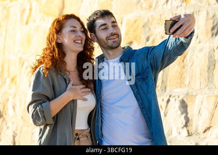 Una giovane coppia sorride calorosamente mentre si fa un selfie insieme davanti a una luminosa parete di pietra, godendosi un momento spensierato con abiti casual. Lifestyl Foto Stock