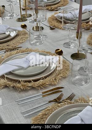 Elegante ambiente da tavolo con tonalità neutre, caratterizzato da tovaglie tessute, posate d'oro, tovaglioli di lino bianco e candele coniche rosa. Foto Stock