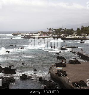 La passeggiata e il lido di puerto de la cruz a tenerife con persone sul lungomare e onde spettacolari che si infrangono sulle rocce costiere Foto Stock