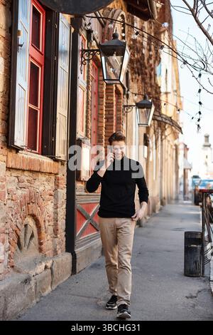 Un uomo elegante cammina lungo una strada pittoresca, chiacchierando al telefono in mezzo a un'architettura unica. Foto Stock