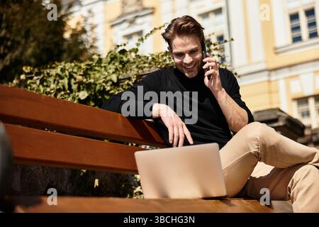 Un uomo elegante con un sorriso sicuro siede su una panchina, chattando al telefono mentre utilizza un laptop. Foto Stock