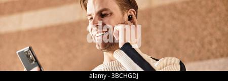 Un bell'uomo passeggia lungo una strada cittadina, sorridendo mentre interagisce con il suo smartphone e ascolta. Foto Stock