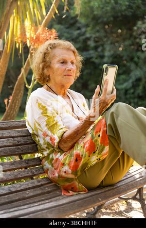 Donna anziana che si gode un momento tranquillo nel parco, seduto comodamente su una panchina mentre naviga sul suo smartphone e si connette con il mondo Foto Stock