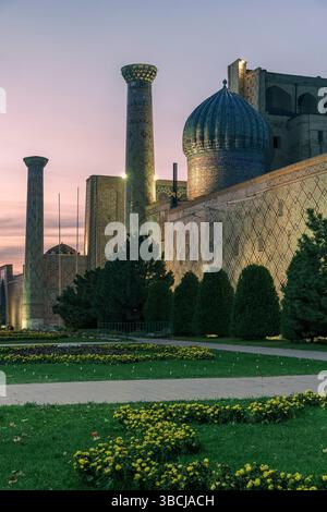 La Madrasa di Ulugh Beg a Registan, Samarcanda, Uzbekistan, è illuminata di notte, con cupole e minareti che si illuminano sopra giardini ben curati. Foto Stock