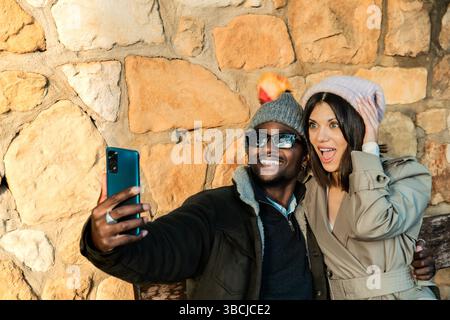 Una coppia allegra vestita con comodi abiti autunnali scatta un selfie mentre si siede su una panchina. L'uomo indossa occhiali da sole e un berretto con un variopinto pom-po Foto Stock