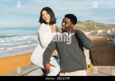 Una coppia gioiosa ride e condivide un momento sincero mentre posa vicino alla spiaggia. Con onde, sabbia e una costa panoramica sullo sfondo, si irradiano Foto Stock