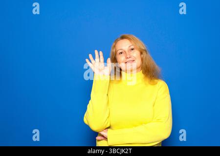 Donna caucasica di mezza età che indossa un maglione giallo su sfondo blu isolato che ondeggia la mano con un'espressione felice Foto Stock