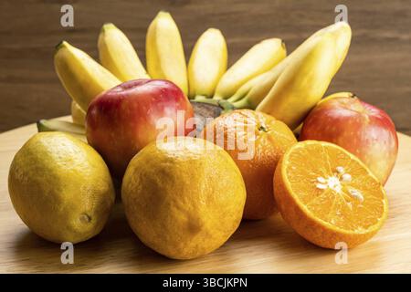 Frutta tropicale matura assortita su tavola di legno. Vari frutti dolci maturi, mazzi di banane, arancia intera e mezza, mele rosse e lemmon maturo Foto Stock