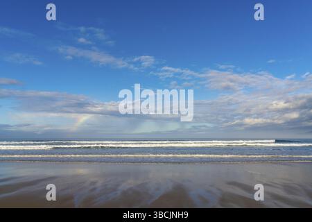 Una spiaggia di sabbia con grandi onde e un piccolo arcobaleno in un cielo espressivo Foto Stock