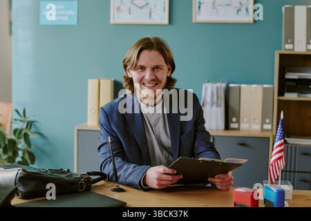 Ritratto di un uomo sorridente che tiene in mano gli appunti mentre è seduto alla scrivania in ufficio con una piccola bandiera americana, un cartello del centro applicazioni e una pianta in vaso Foto Stock