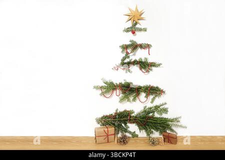 Albero di Natale fai da te realizzato con rami naturali inchiodati a una parete bianca, catena decorativa rossa e una stella di carta in cima, regali sul pavimento in legno, semplice Foto Stock