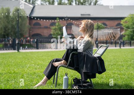 Donna scomoda che si sventola con fogli di carta come un ventilatore mentre si siede sulla sedia all'esterno sul prato. Foto Stock
