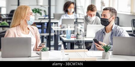 Lavoro di squadra e tecnologia moderna. Giovane uomo e donna in maschere protettive con smartphone in mano sul posto di lavoro con computer portatili e antisettici, che parla Foto Stock