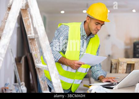 Man engineer che controlla i documenti e utilizza il computer portatile durante gli interventi di riparazione Foto Stock
