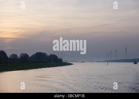 Turbine eoliche e navi fiancheggiano il fiume Nieuwe Maas al tramonto vicino a Rotterdam, che si staglia contro i cieli pastello e la morbida foschia industriale. Foto Stock