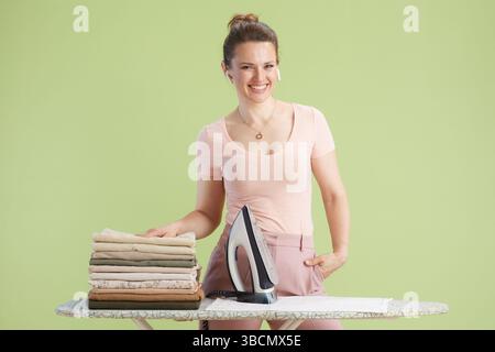Una donna sorridente sta dietro un asse da stiro con le mani in tasca, creando un'atmosfera rilassata. Le tonalità del rosa chiaro e del verde oliva esaltano il Foto Stock