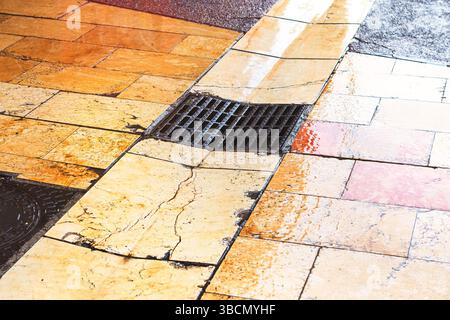 Passerella piastrellata con griglia di drenaggio e coperchio per tombino. Le piastrelle sono bagnate, a indicare la pioggia recente e alcune sono incrinate e irregolari. L'acqua fluisce attraverso la p Foto Stock