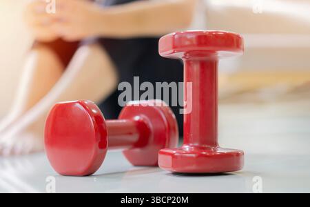 Due manubri rossi sul pavimento con l'immagine sfocata di una donna stanca e stanca in un abbigliamento sportivo che riposa dietro. Messa a fuoco selettiva. Foto Stock