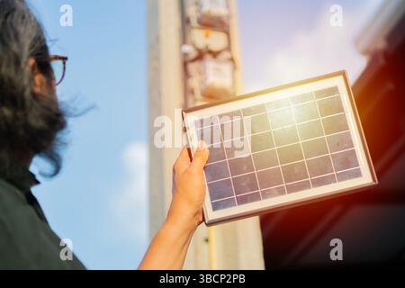 Uomo maturo con mini pannello a celle solari con sfondo sfocato di palo in cemento con misuratore elettrico. Messa a fuoco selettiva. Foto Stock