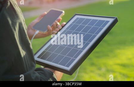 Tecnologie moderne di energia portatile. Colpo di uomo che tiene il pannello solare e il telefono in piedi sull'erba sotto i caldi e morbidi raggi del sole Foto Stock