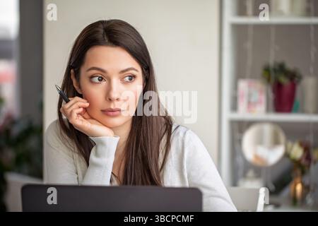 Donna premurosa con penna seduta al computer portatile in un luminoso ufficio domestico Foto Stock
